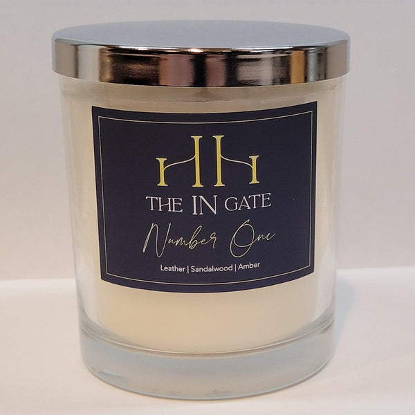 The In Gate Candle Collection. Number One. Clear glass candle with navy and yellow label, and a shiny silver lid.