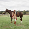 A woman in the Welligogs Damelza Camel Coat, white pants, and brown boots stands beside a dark brown horse in an open grassy field under an overcast sky.