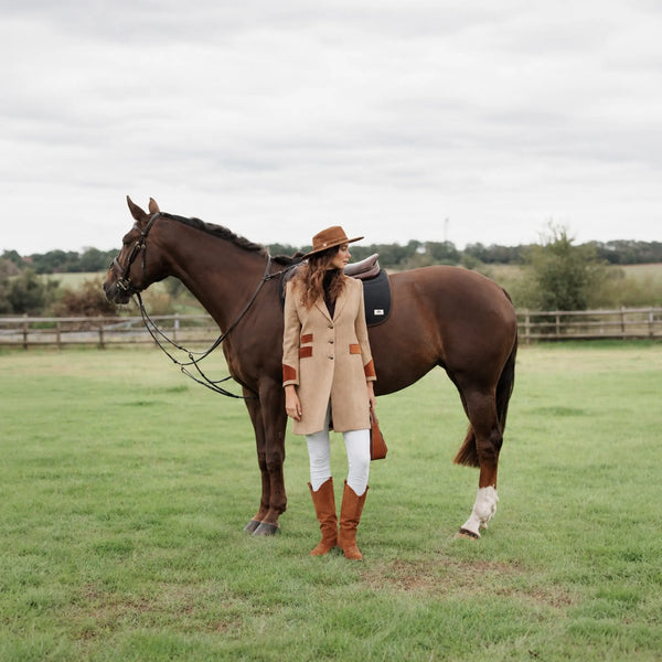 A woman in the Welligogs Damelza Camel Coat, white pants, and brown boots stands beside a dark brown horse in an open grassy field under an overcast sky.