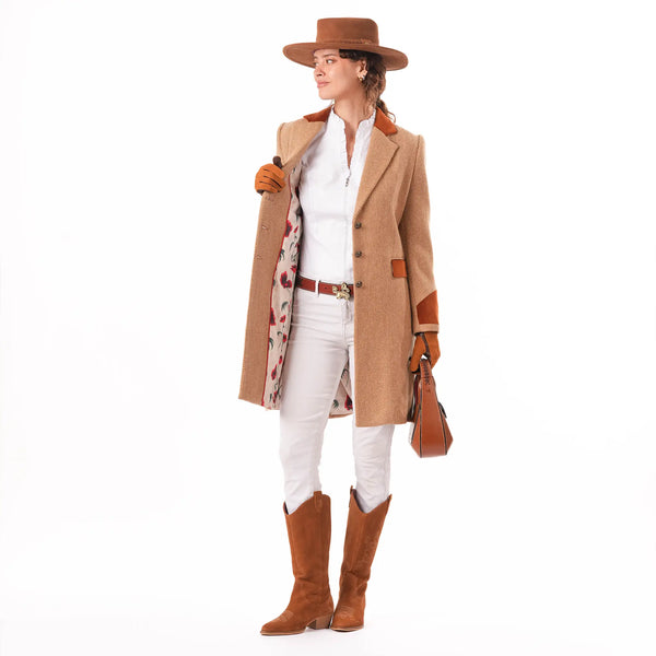A woman modeling the Welligogs Damelza Camel Coat - a mid-length camel coat with brown leather trim and a poppy floral lining, styled with a brown hat, white shirt and pants, brown boots, and carrying a brown handbag against a white background.