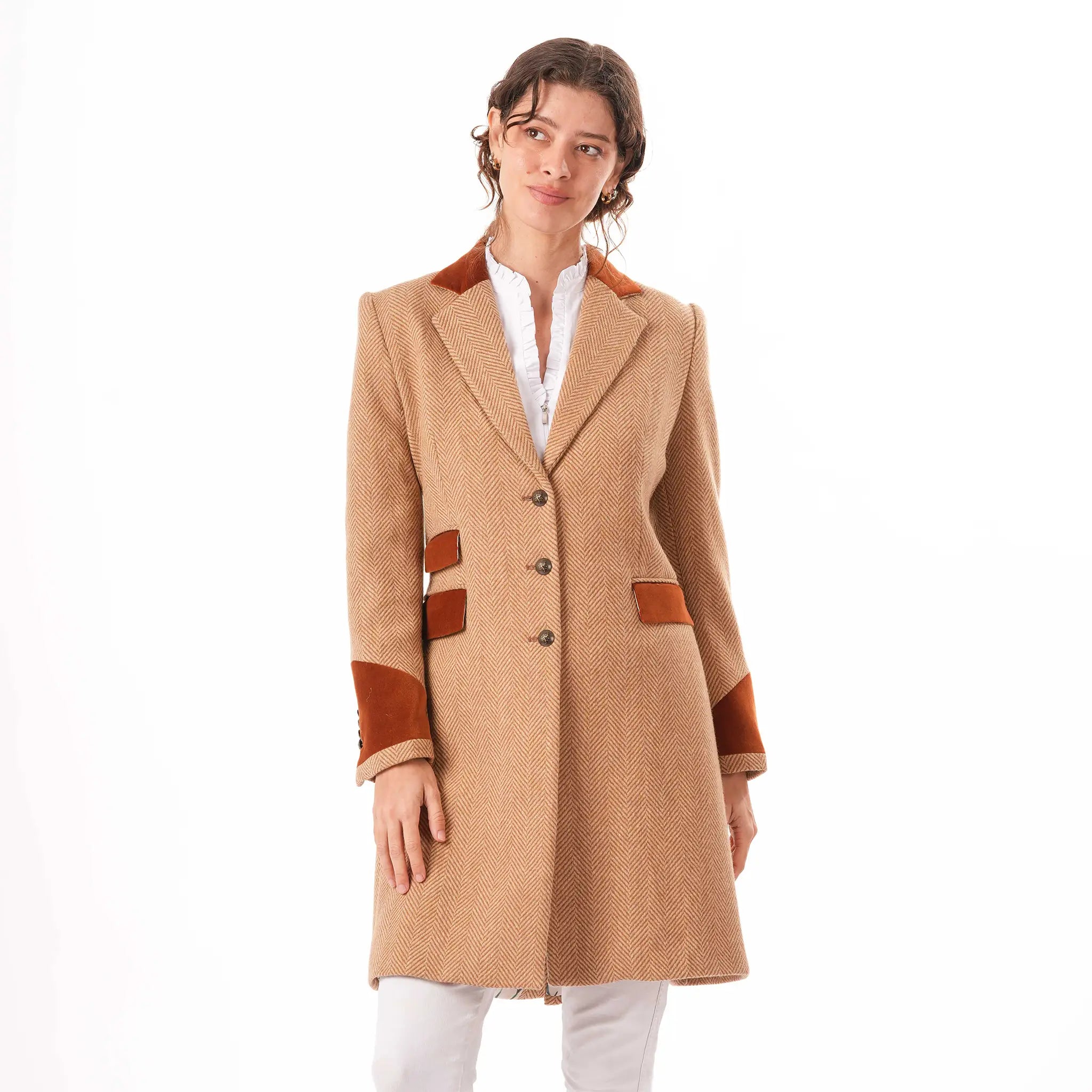 A woman modeling the Welligogs Damelza Camel Coat - a wool coat with brown suede-like trim on the collar, cuffs, and pockets, worn over a white blouse and white trousers against a plain white background.