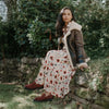 Woman in a floral dress and brown coat sitting outdoors among greenery