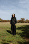 A woman stands in a grassy field under a blue sky, wearing a black waxed cotton jacket and black wide-leg trousers.