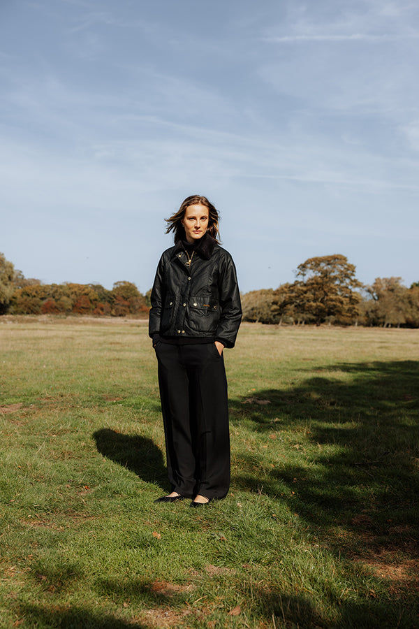 A woman stands in a grassy field under a blue sky, wearing a black waxed cotton jacket and black wide-leg trousers.