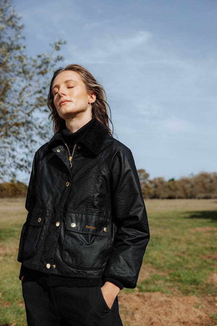 A woman wearing a black waxed cotton jacket with gold-tone snap buttons and a black corduroy collar, standing outdoors in a grassy field under a blue sky.