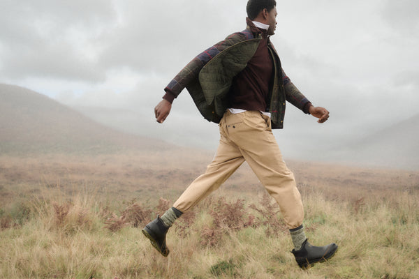 A man walking through a grassy, misty outdoor landscape wearing a quilted olive green jacket, a brown sweater, beige trousers, and black boots.