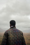 A man viewed from behind wearing a quilted plaid jacket in an outdoor landscape under a cloudy sky.