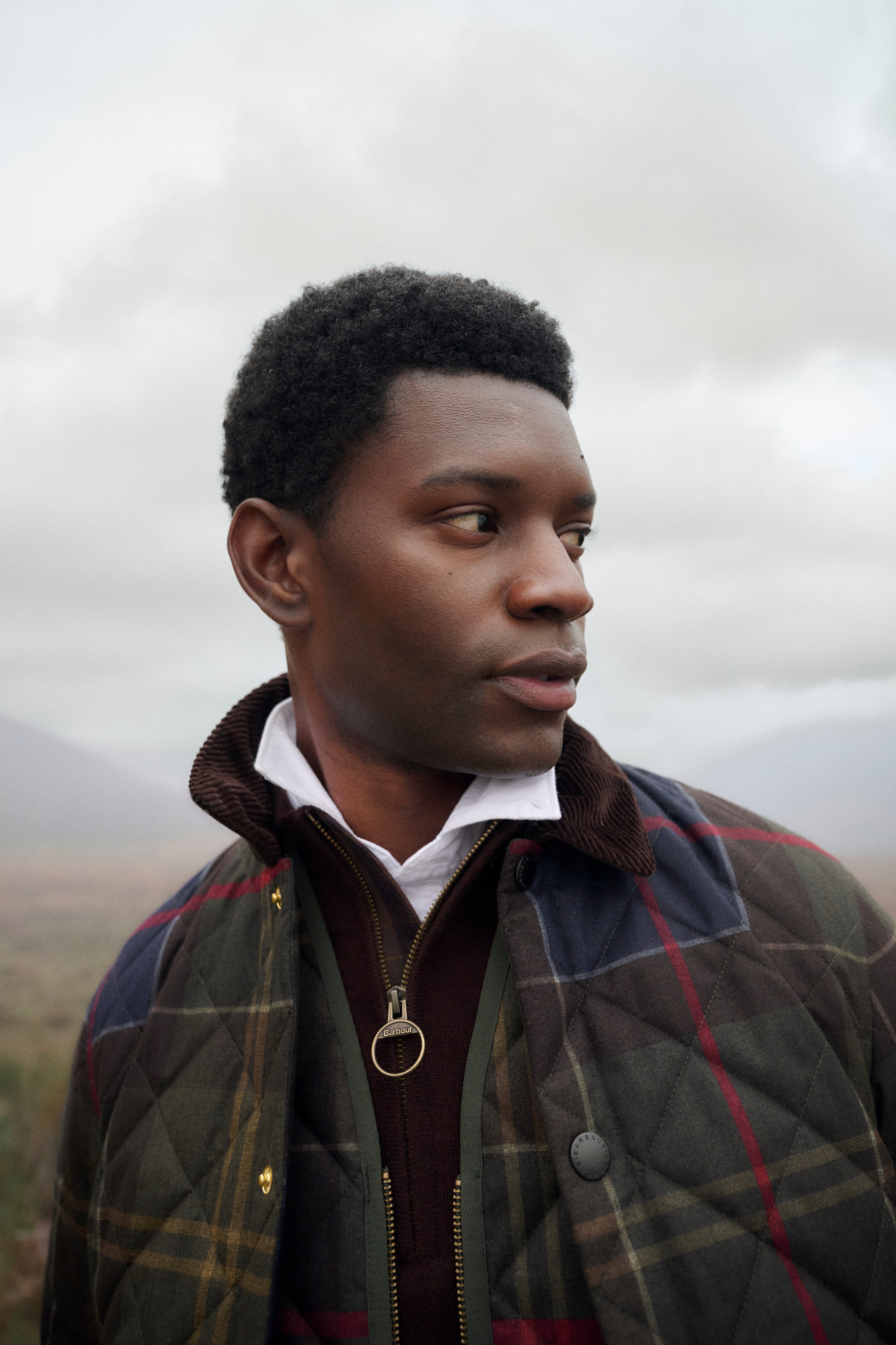 A man in profile wearing a quilted plaid jacket over a white collared shirt and dark zip-up layer, photographed outdoors against a cloudy sky and distant hills.