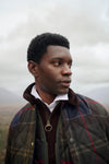 A man in profile wearing a quilted plaid jacket over a white collared shirt and dark zip-up layer, photographed outdoors against a cloudy sky and distant hills.