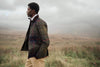 A man in profile wearing a quilted plaid jacket and beige trousers standing in a misty grassy field.
