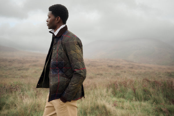 A man in profile wearing a quilted plaid jacket and beige trousers standing in a misty grassy field.