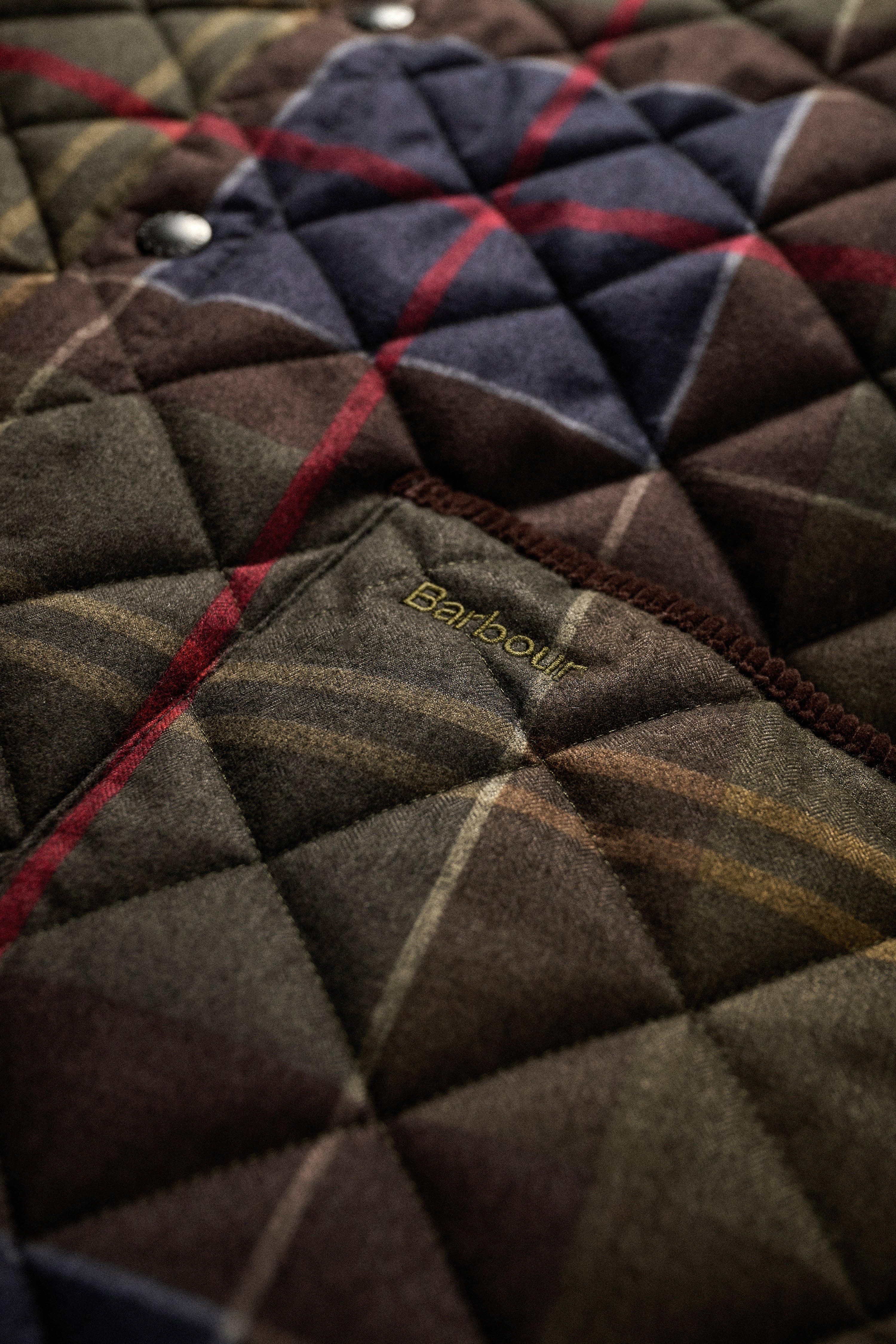 Close-up of a quilted Barbour jacket fabric with a diamond-stitched pattern, featuring a plaid design in dark brown, navy, red, and beige, with the Barbour logo embroidered on a pocket.