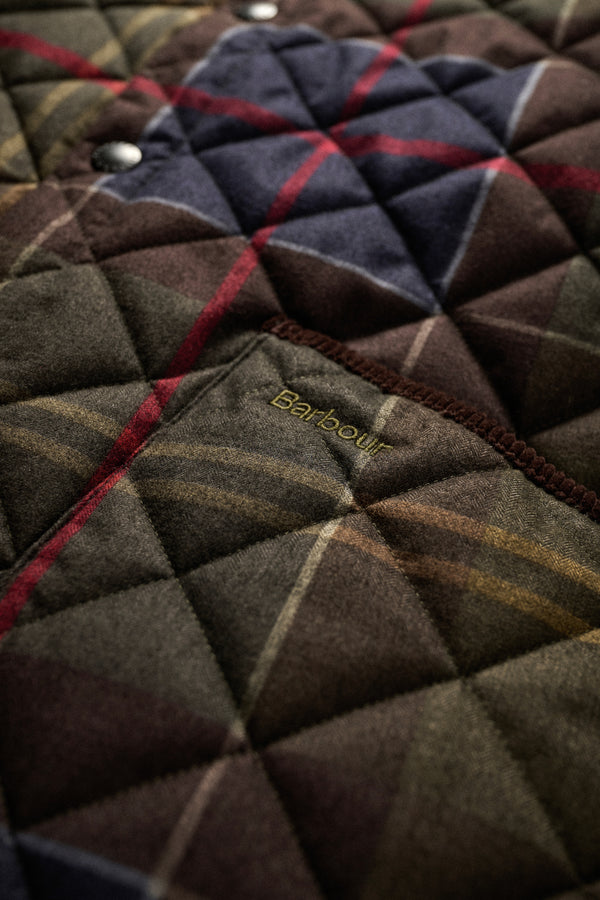 Close-up of a quilted Barbour jacket fabric with a diamond-stitched pattern, featuring a plaid design in dark brown, navy, red, and beige, with the Barbour logo embroidered on a pocket.