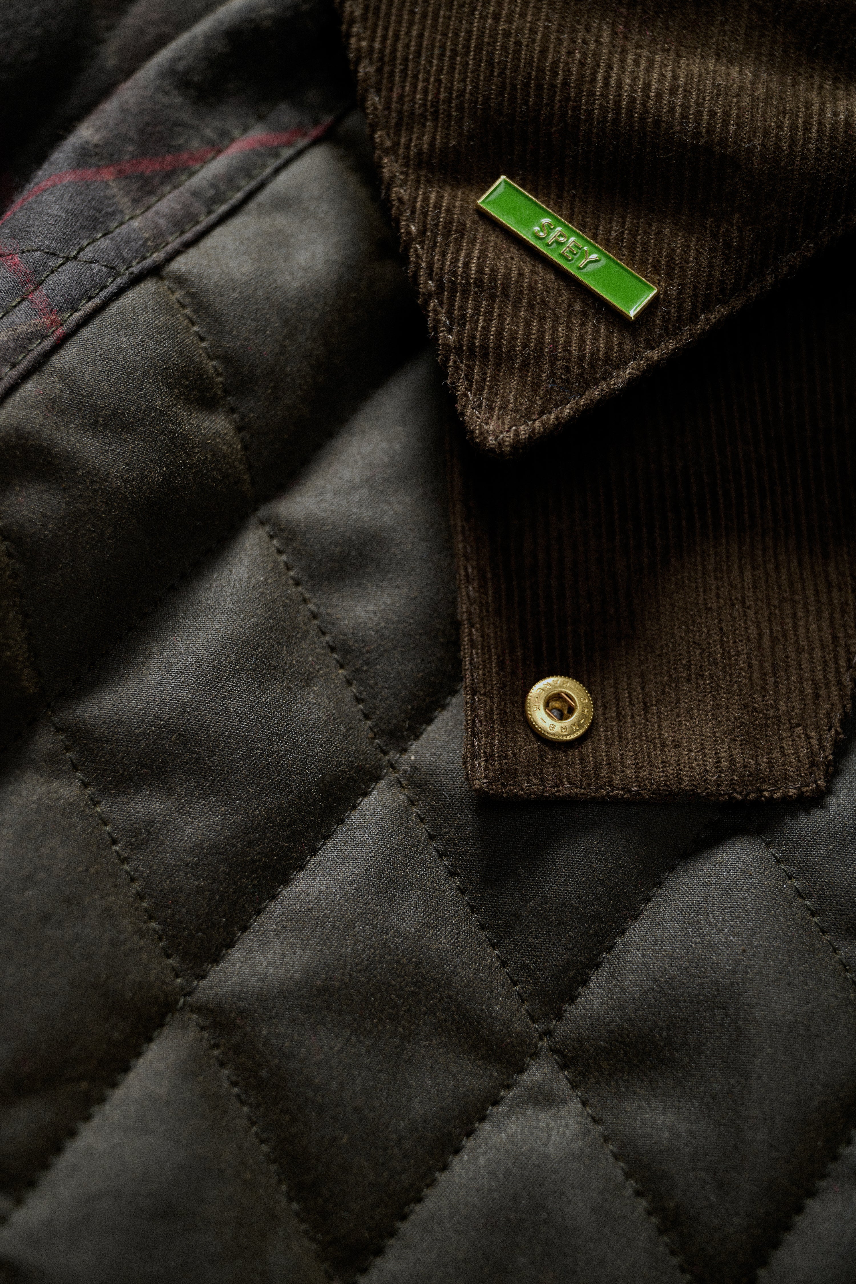 Close-up of a dark brown corduroy collar with a gold snap button and a small green brand tag reading 'SPEY', next to a dark quilted fabric panel.