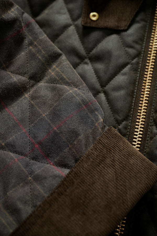Close-up detail of a quilted jacket showing dark olive and brown plaid fabric, a dark brown corduroy collar, and a gold-toned zipper.