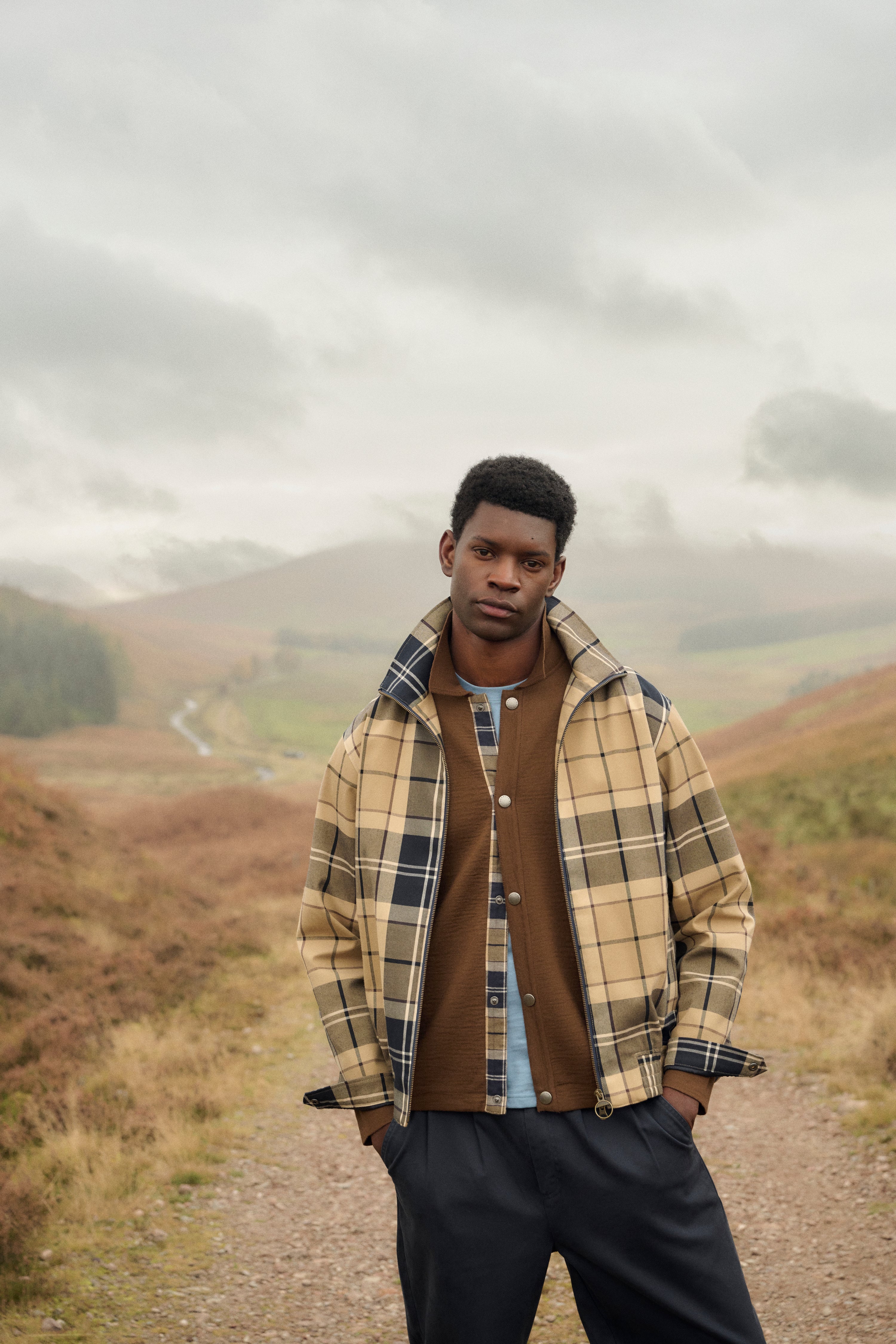 Man wearing a Barbour Tartan Showerproof Blouson Jacket in a scenic outdoor setting with mountains and a cloudy sky.