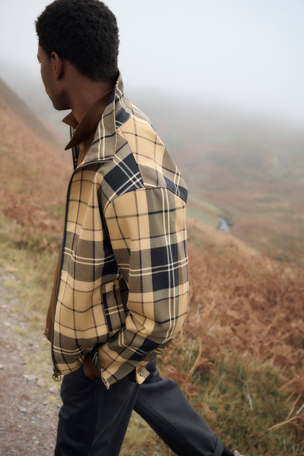 A man wearing a beige and navy Barbour Tartan Showerproof Blouson Jacket with a brown collar, walking along a path in a misty, grassy outdoor landscape.