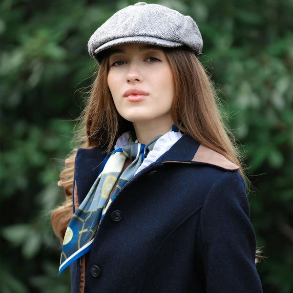 Woman wearing a navy coat, gray cap, and patterned scarf with a blurred green background