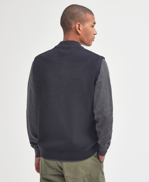 Man wearing a dark gray sweater vest over a gray shirt with green pants on a light gray background