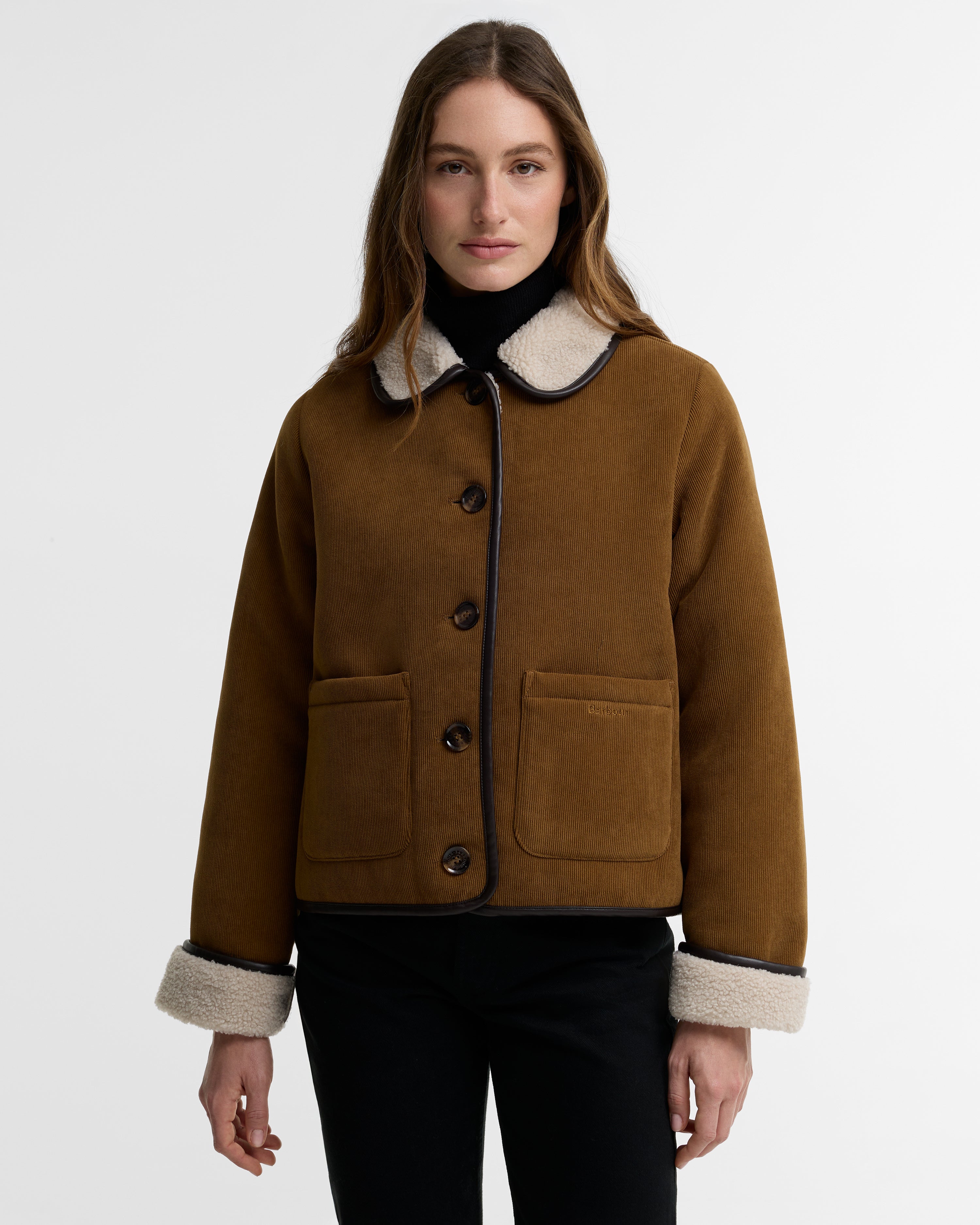 A woman modeling a brown suede shearling jacket with a cream-colored collar and cuffs, paired with a black turtleneck and black pants, against a plain light gray background.