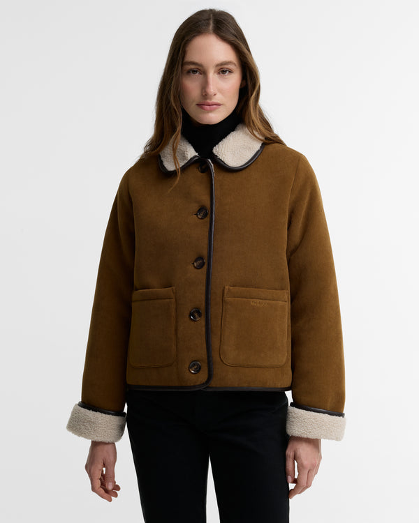 A woman modeling a brown suede shearling jacket with a cream-colored collar and cuffs, paired with a black turtleneck and black pants, against a plain light gray background.