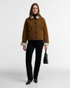 A model stands against a light gray studio background wearing a cropped brown shearling jacket with a cream collar and cuffs, black trousers, black shoes, and holding a black handbag.