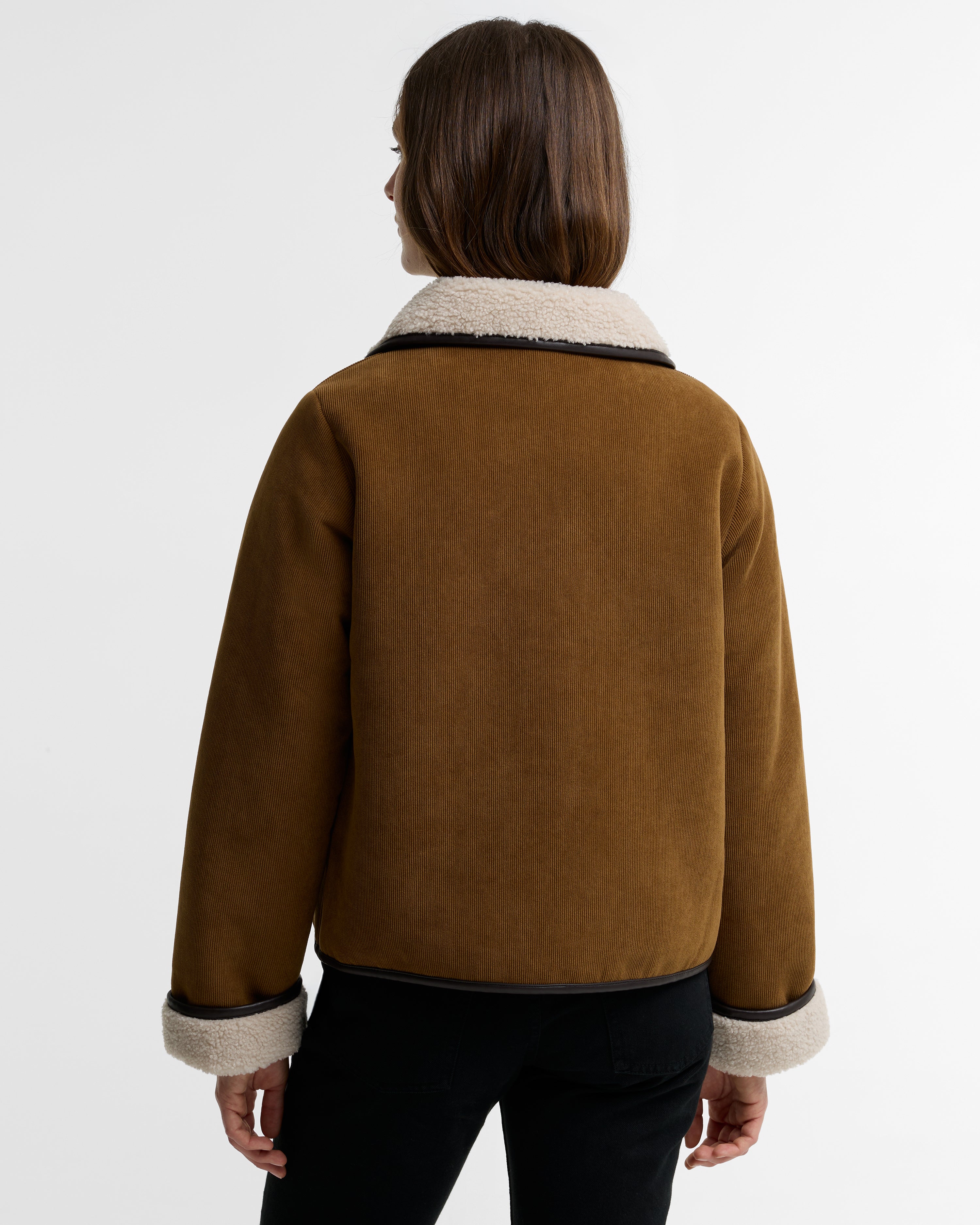 A woman viewed from the back wearing a brown suede-like jacket with a cream shearling-style collar and cuffs, paired with black pants, against a plain light gray studio background.