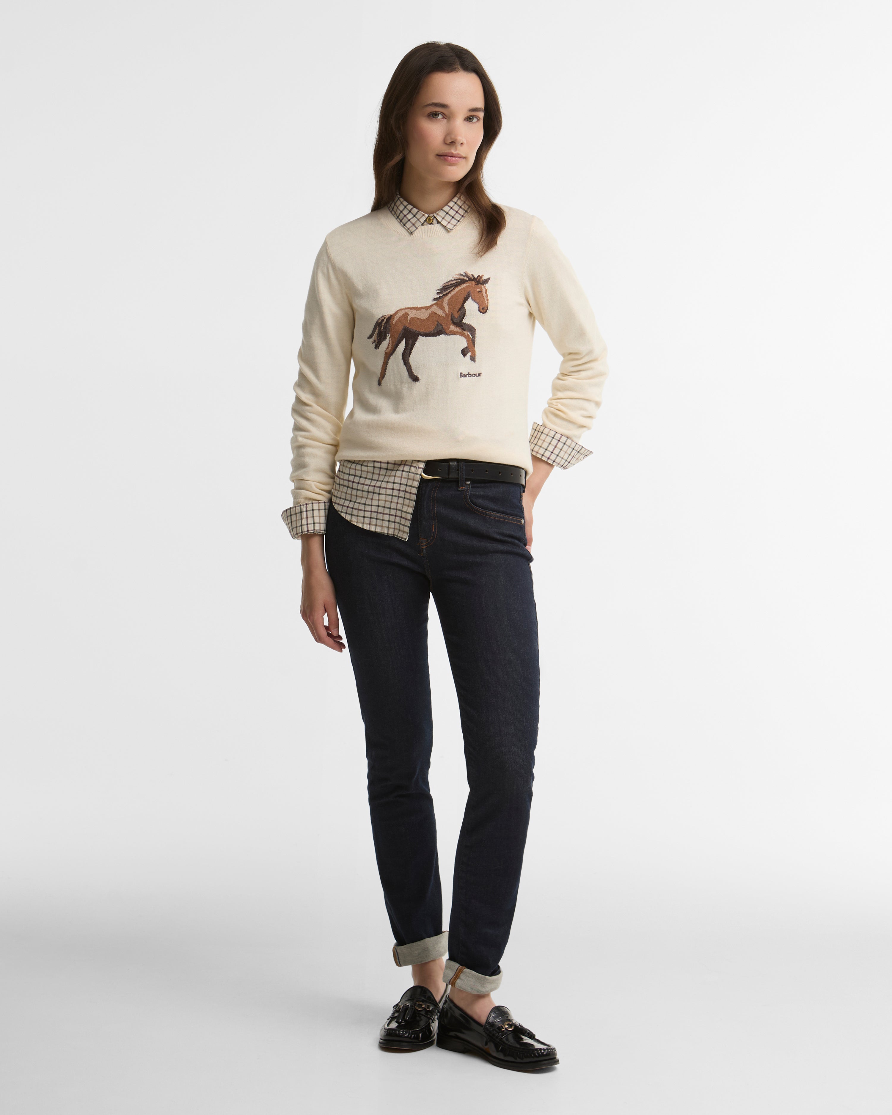 Woman wearing a cream sweater with a horse design, checkered shirt, and black pants on a white background