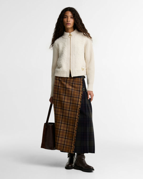 Model wearing the Barbour Hazelwood Knitted Zip Through Jacket in cream, styled with a brown tartan maxi skirt, black boots, and a dark leather tote bag.