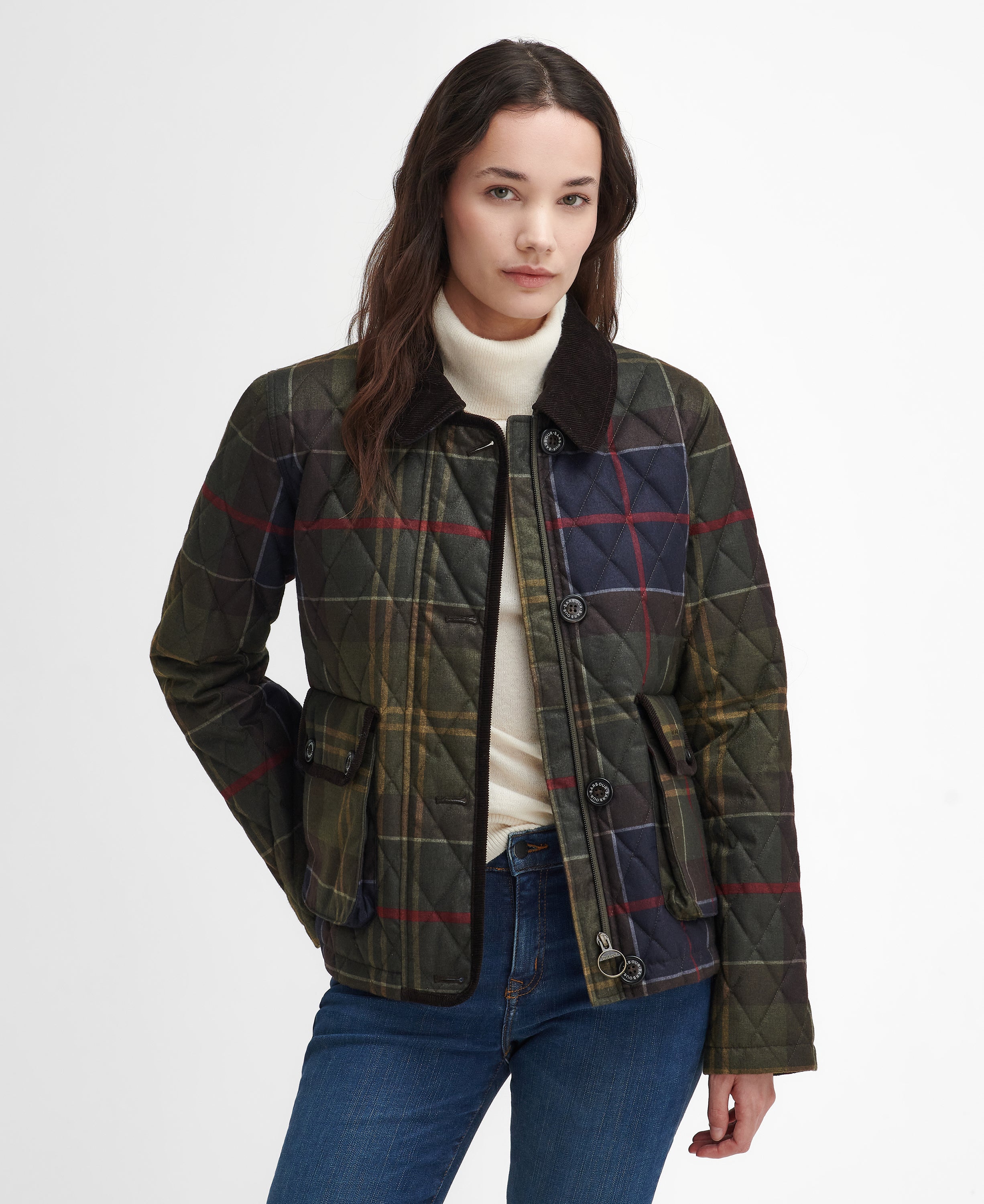 A woman modeling a quilted plaid jacket with a dark brown collar, worn over a cream turtleneck and blue jeans, against a plain white studio background.