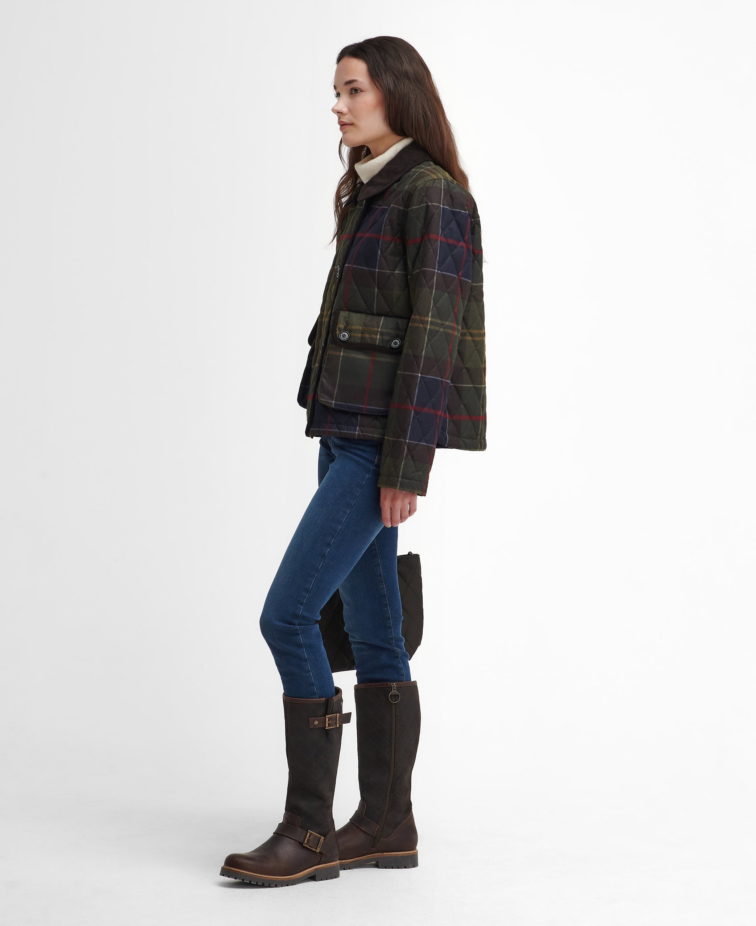 A woman stands in profile wearing a dark tartan plaid jacket with red and yellow lines, blue jeans, and dark brown knee-high boots against a plain white studio background.