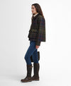 A woman stands in profile wearing a dark tartan plaid jacket with red and yellow lines, blue jeans, and dark brown knee-high boots against a plain white studio background.