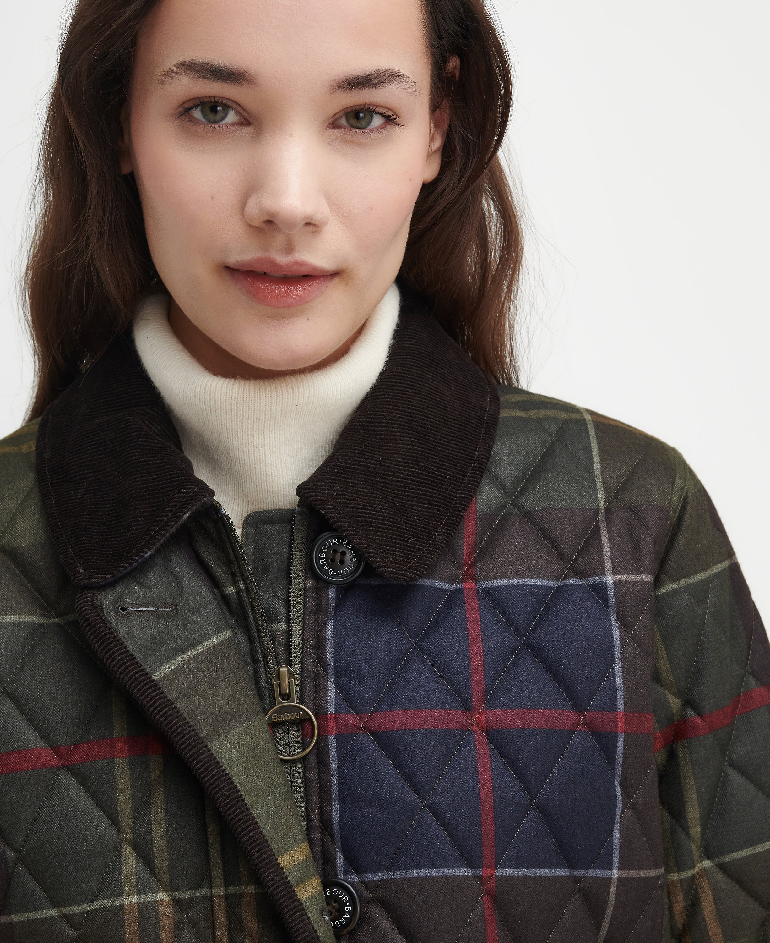 Close-up of a woman wearing a quilted plaid jacket with a dark brown corduroy collar over a cream turtleneck, against a plain light background.