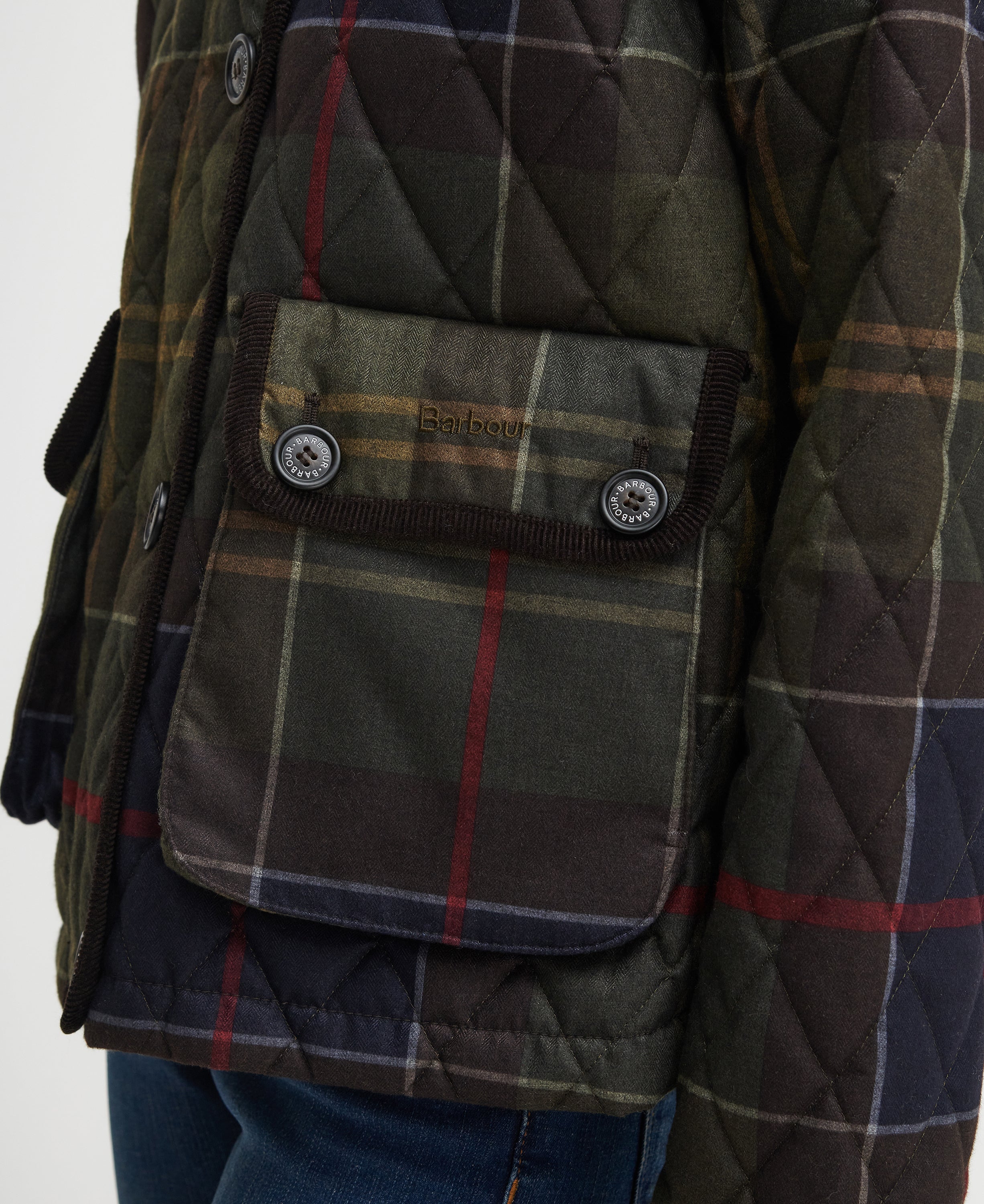 Close-up of a quilted Barbour jacket in a green and brown plaid pattern, featuring a front flap pocket with snap buttons and the Barbour logo.