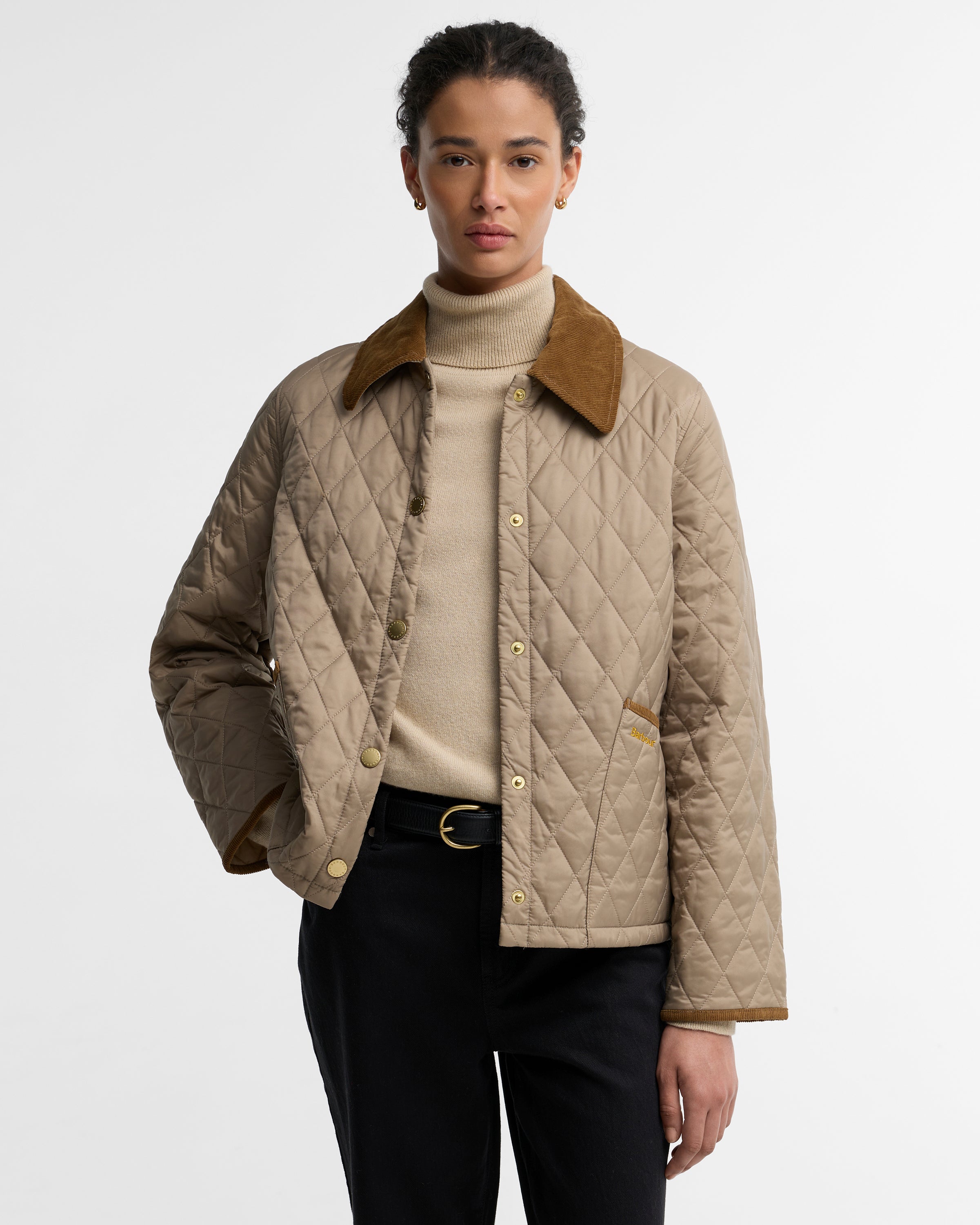 A model wearing a beige diamond-quilted jacket with a brown corduroy collar, layered over a beige turtleneck sweater and black trousers, shot against a plain light gray background.