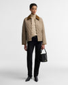A model stands against a light gray studio background wearing a beige quilted jacket with a brown collar, a cream turtleneck, black trousers, black pointed shoes, and holding a small black handbag.