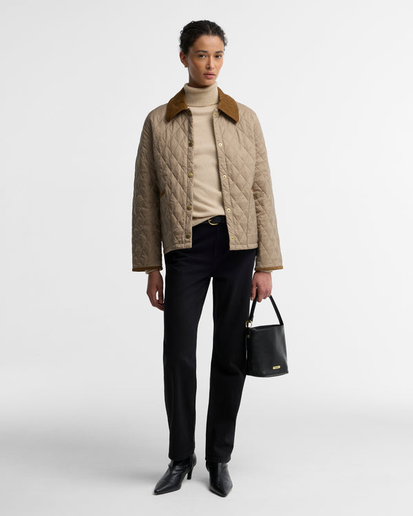 A model stands against a light gray studio background wearing a beige quilted jacket with a brown collar, a cream turtleneck, black trousers, black pointed shoes, and holding a small black handbag.