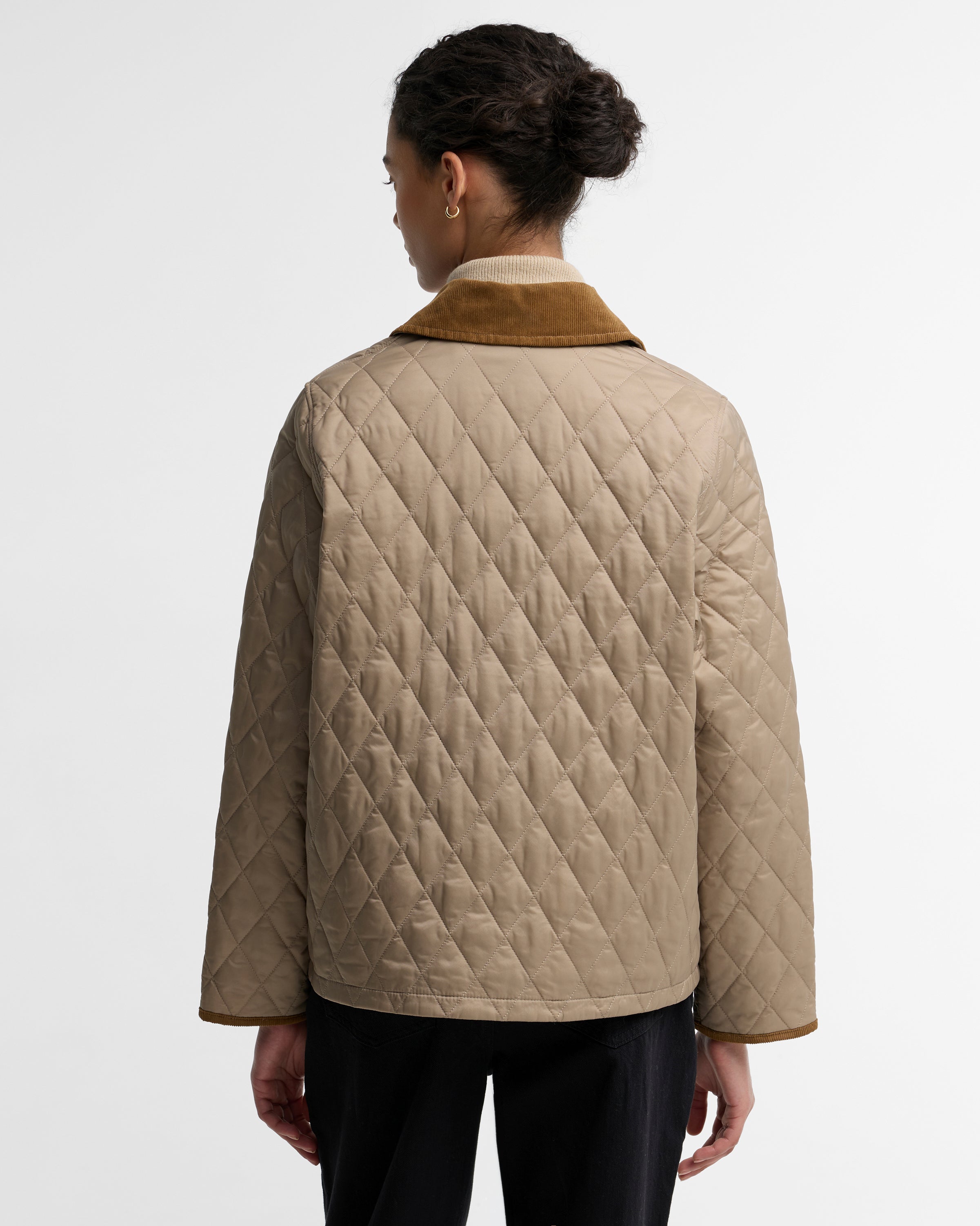 A woman shown from the back wearing a beige quilted jacket with a brown corduroy collar, paired with black pants, against a plain light gray studio background.