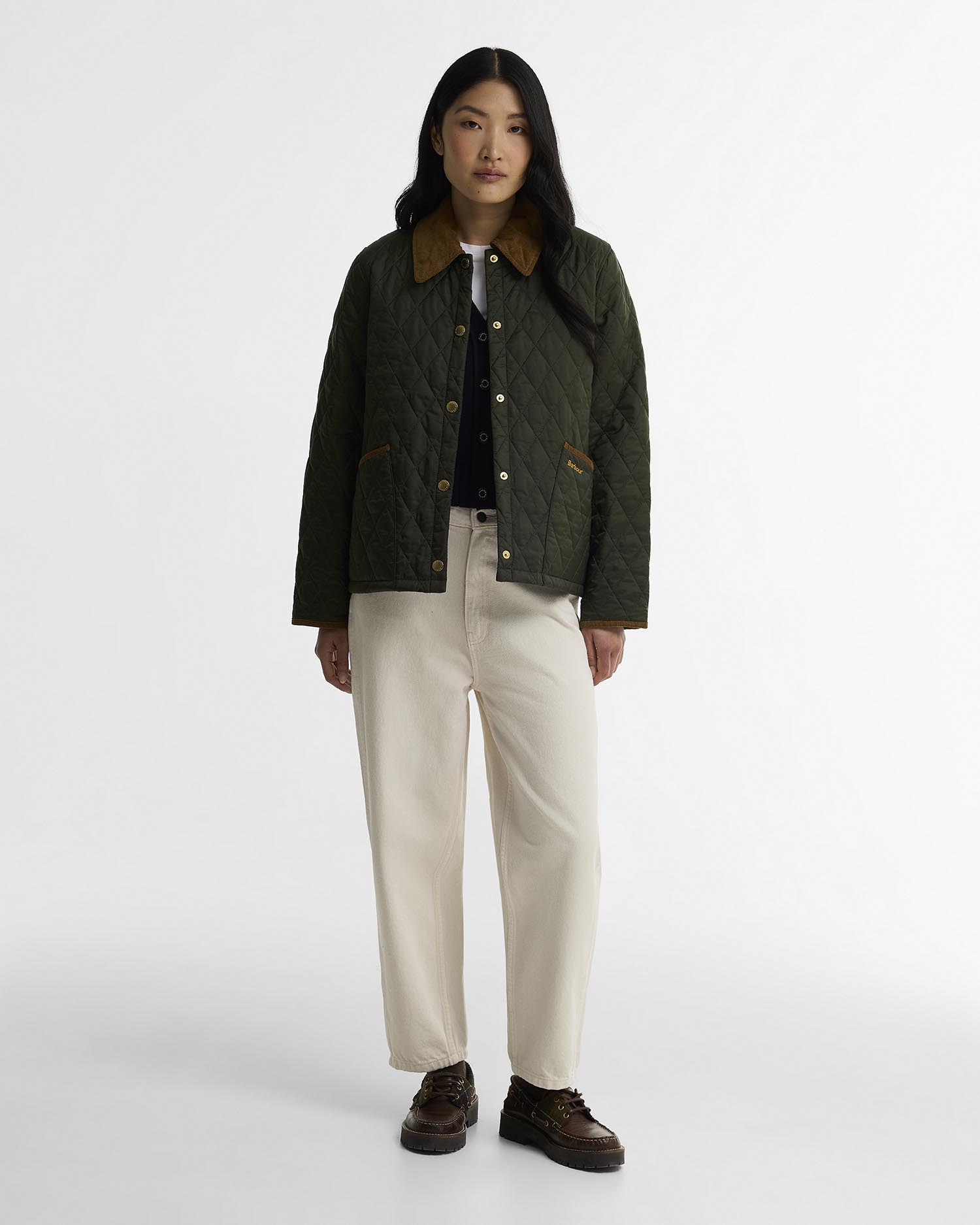 A model stands against a light gray studio background wearing a dark olive green quilted jacket with a brown collar, cream wide-leg trousers, and dark brown shoes.