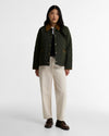 A model stands against a light gray studio background wearing a dark olive green quilted jacket with a brown collar, cream wide-leg trousers, and dark brown shoes.