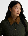 A close-up of a woman wearing a dark olive green quilted jacket with a contrasting brown suede-like collar and gold-tone buttons, set against a plain light gray background.