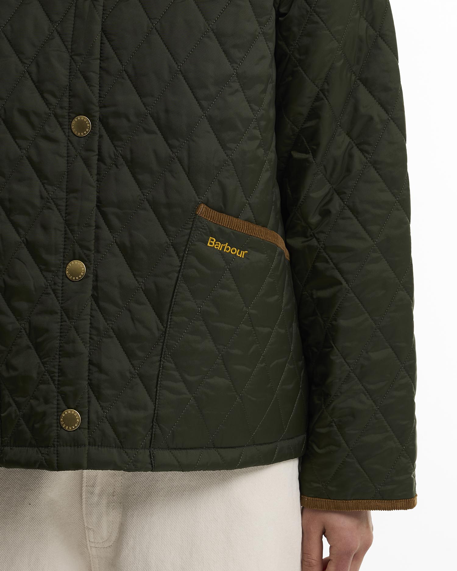Close-up of a quilted Barbour jacket in dark olive green with brass buttons, a brown pocket trim, and gold Barbour embroidery on the pocket, worn by a model.