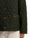 Close-up of a quilted Barbour jacket in dark olive green with brass buttons, a brown pocket trim, and gold Barbour embroidery on the pocket, worn by a model.