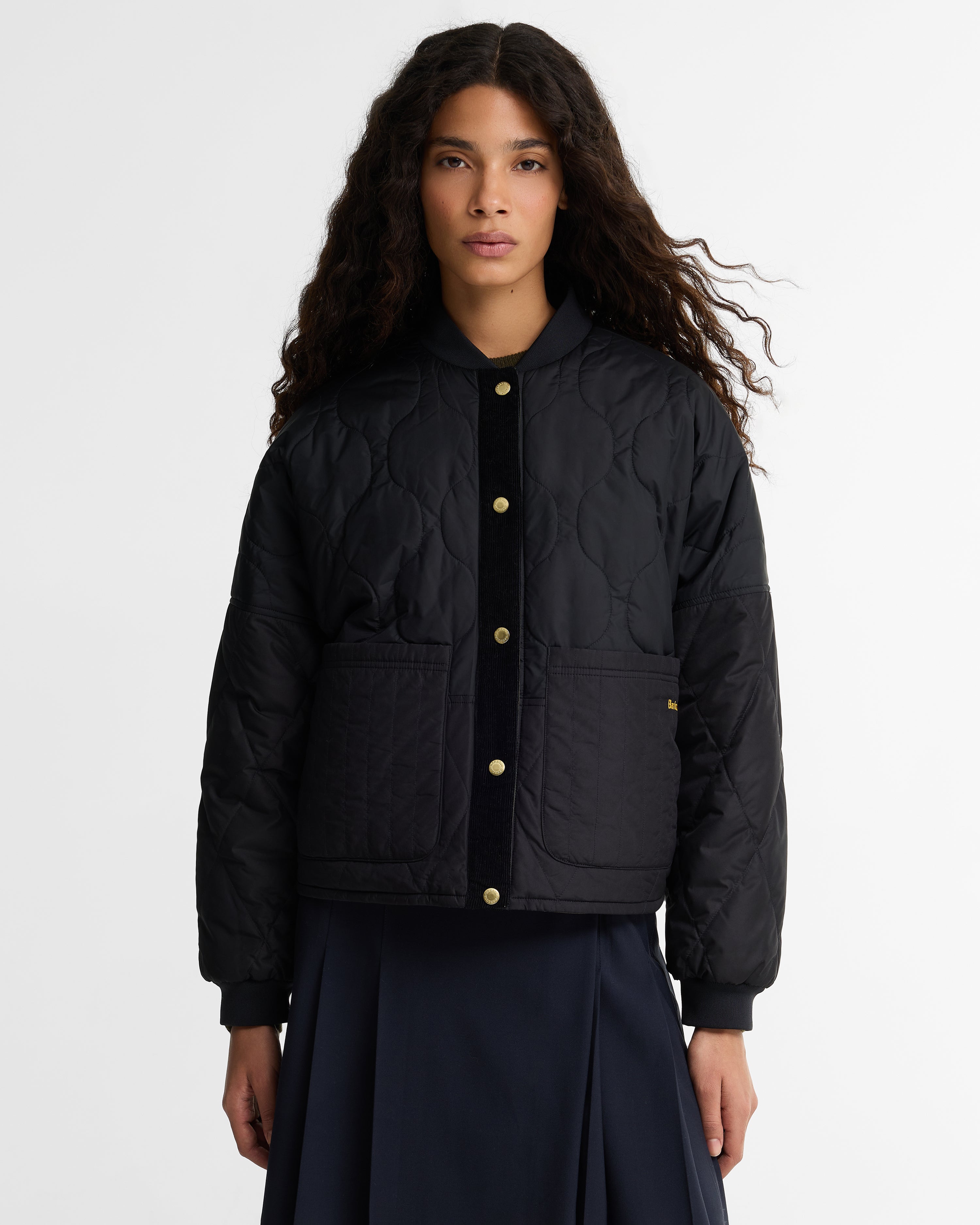 Model wearing the Barbour Ravenshill Reversible Quilted Jacket black-side out, with gold snap buttons, ribbed hem and cuffs, and gold Barbour script on the  pocket, styled with a navy pleated skirt.
