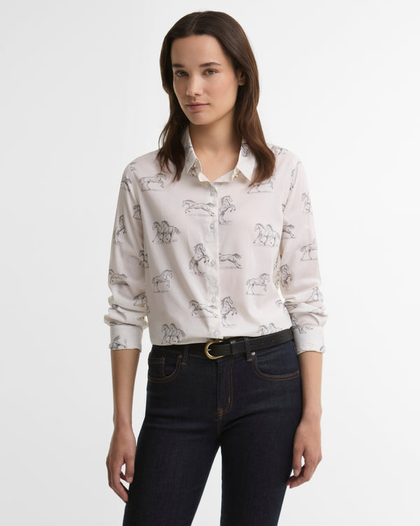 A woman modeling a long-sleeve button-up shirt with a delicate horse pattern, paired with dark blue jeans and a black belt, against a plain white studio background.