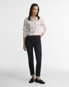 A woman modeling a light-colored, long-sleeve button-up shirt with a delicate pattern of horses, paired with dark wash jeans, black loafers, and a gold belt, set against a plain light gray studio background.