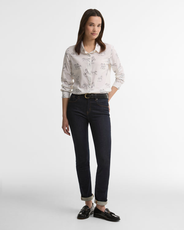 A woman modeling a light-colored, long-sleeve button-up shirt with a delicate pattern of horses, paired with dark wash jeans, black loafers, and a gold belt, set against a plain light gray studio background.