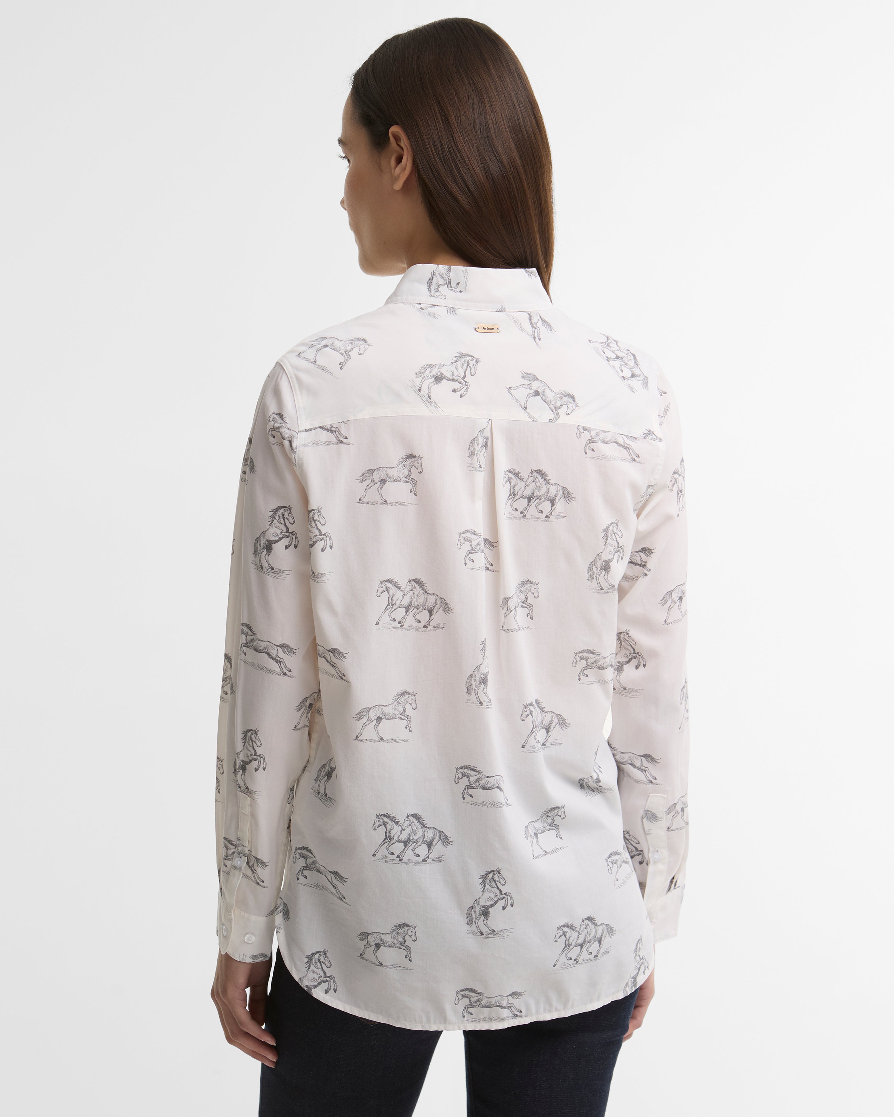 A woman viewed from the back wearing a white long-sleeve button-up shirt with a black horse line-art pattern, paired with dark pants, against a plain light gray studio background.