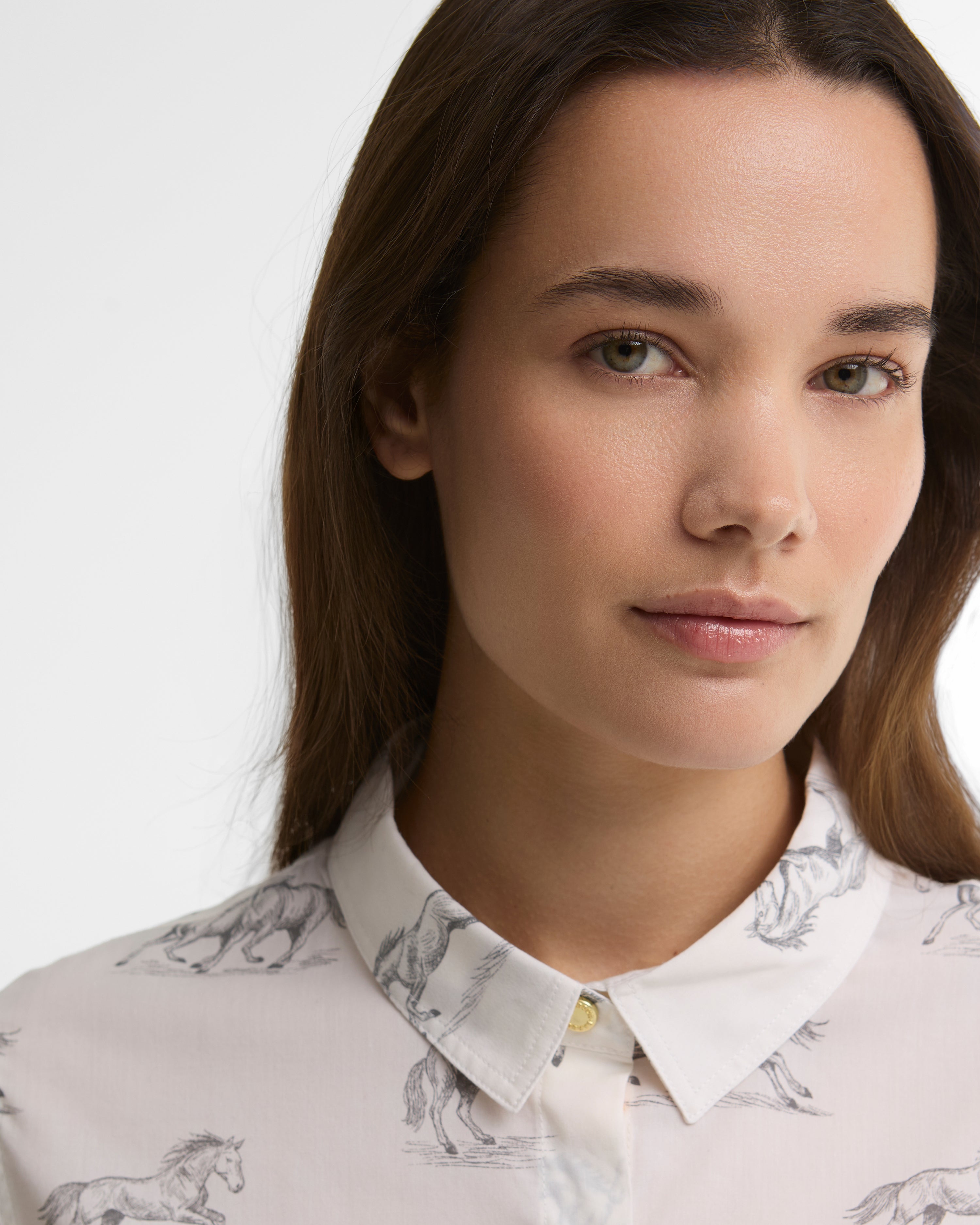 Close-up of a woman wearing a white collared button-up shirt with a subtle gray horse pattern, set against a plain light gray background.