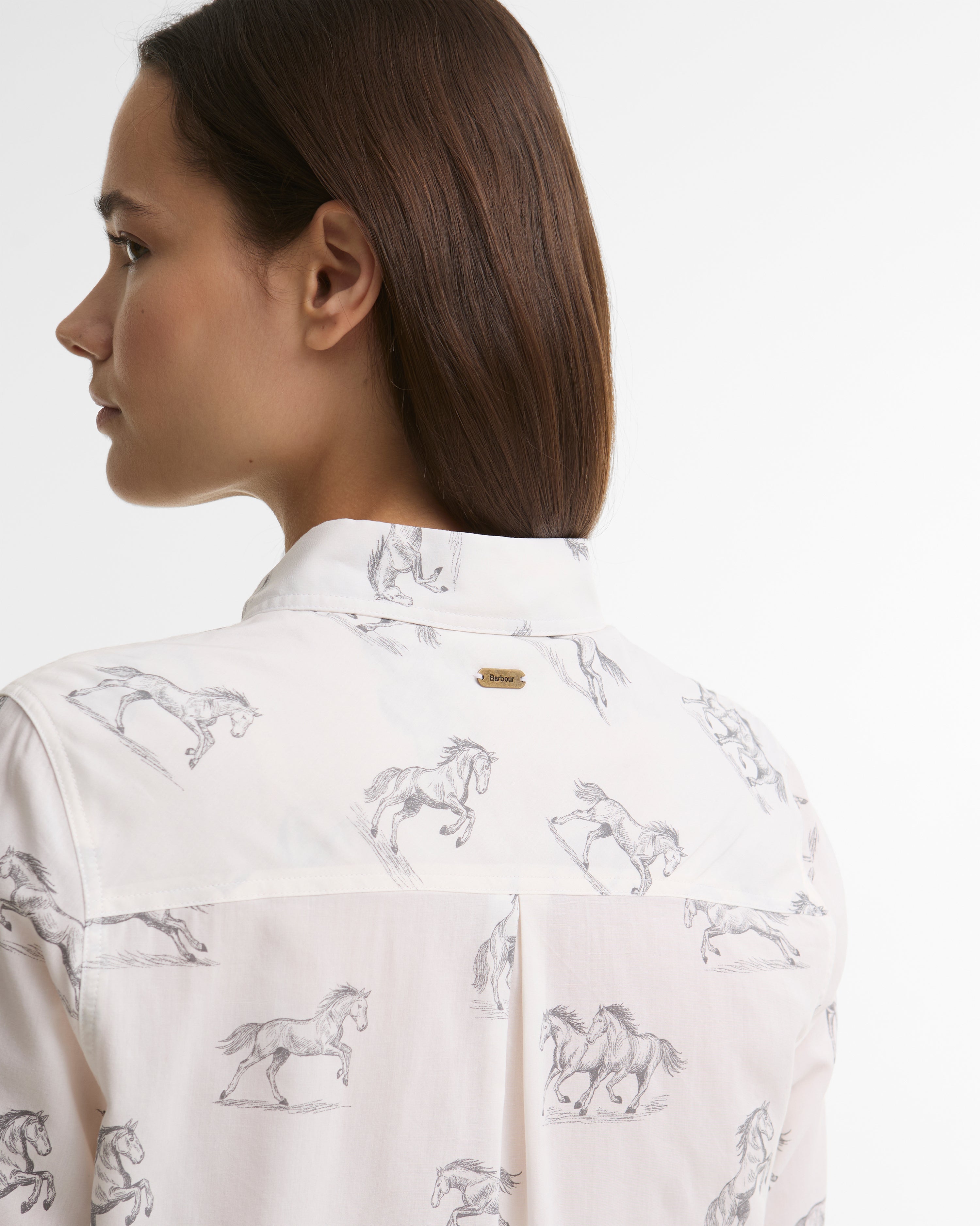 A woman shown from the back wearing a white button-up shirt with a gray horse pattern and a small gold Barbour brand plaque on the collar.